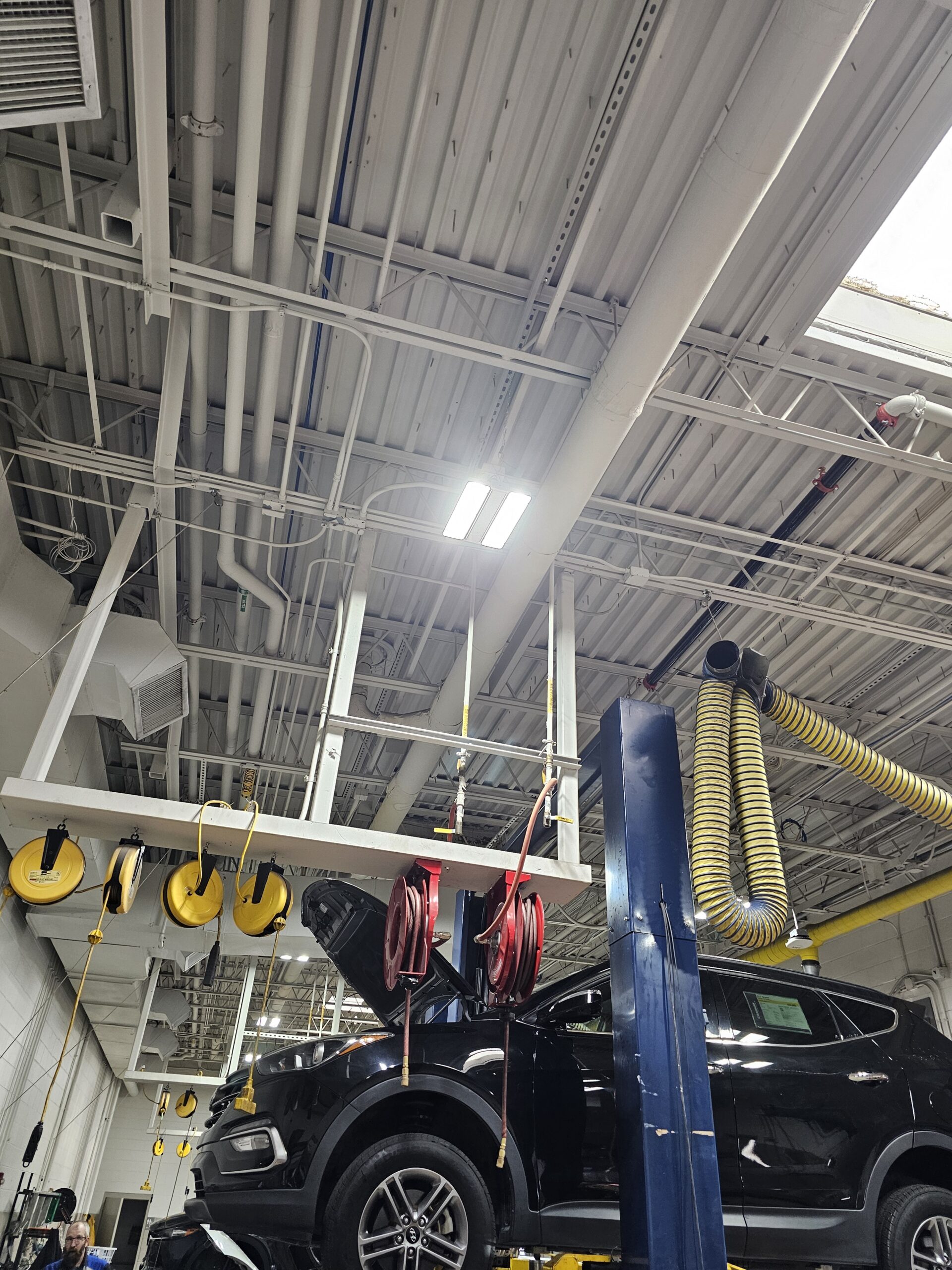 A black SUV is lifted on a blue hydraulic car lift in an auto repair shop, with hoses and cables hanging from the ceiling.