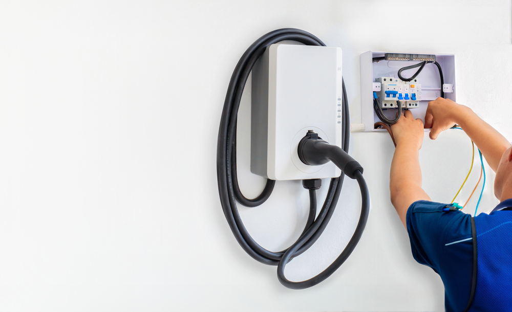 Person installing or servicing an electric vehicle charging station on a white wall, with cables connected to a control panel.