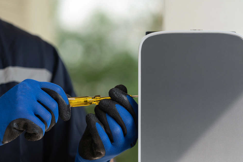 A person wearing blue gloves uses a screwdriver to install or repair a gray electronic device mounted on a wall.