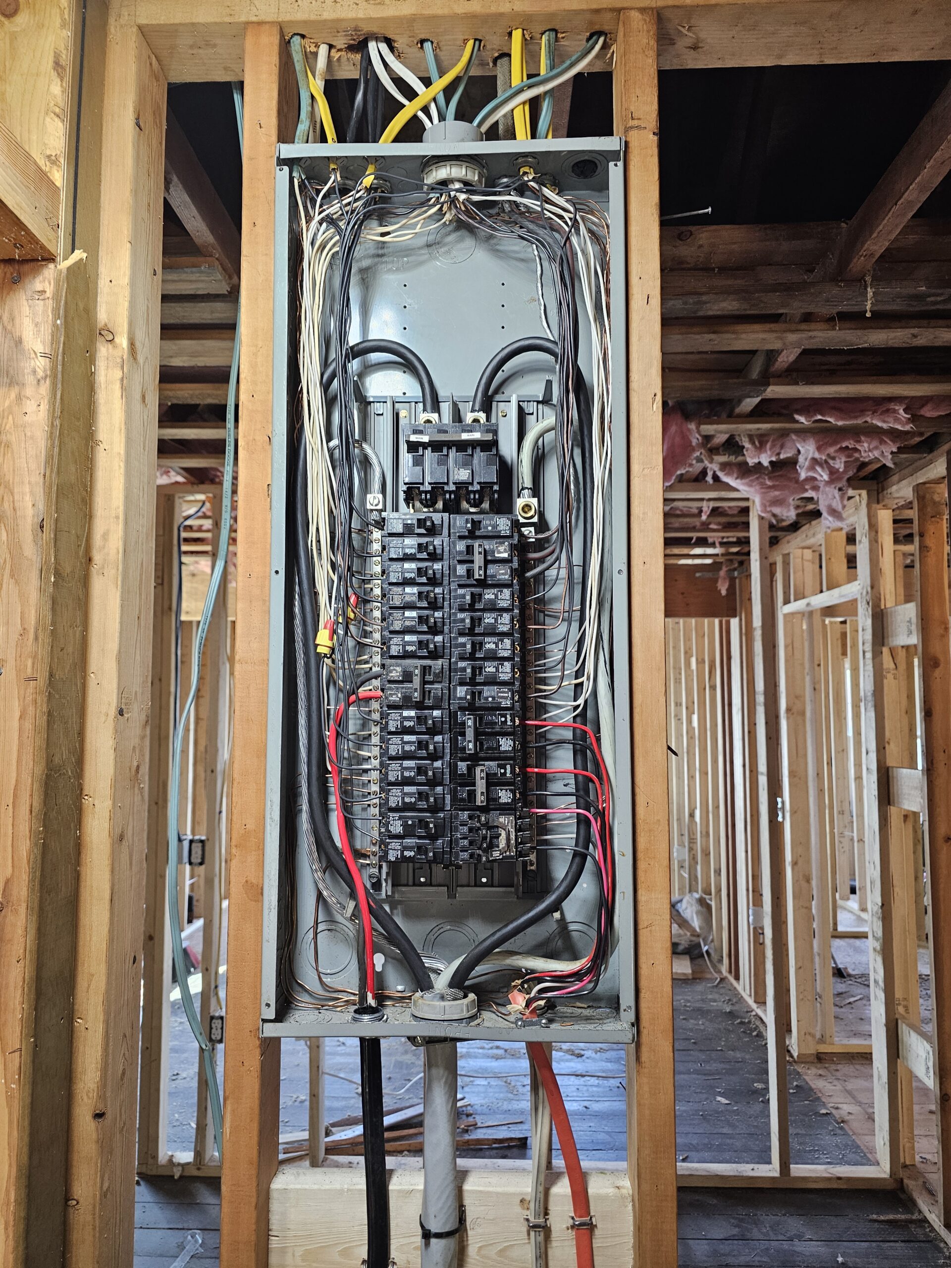 An open electrical panel with exposed wiring is mounted between wooden wall studs in a house under construction.