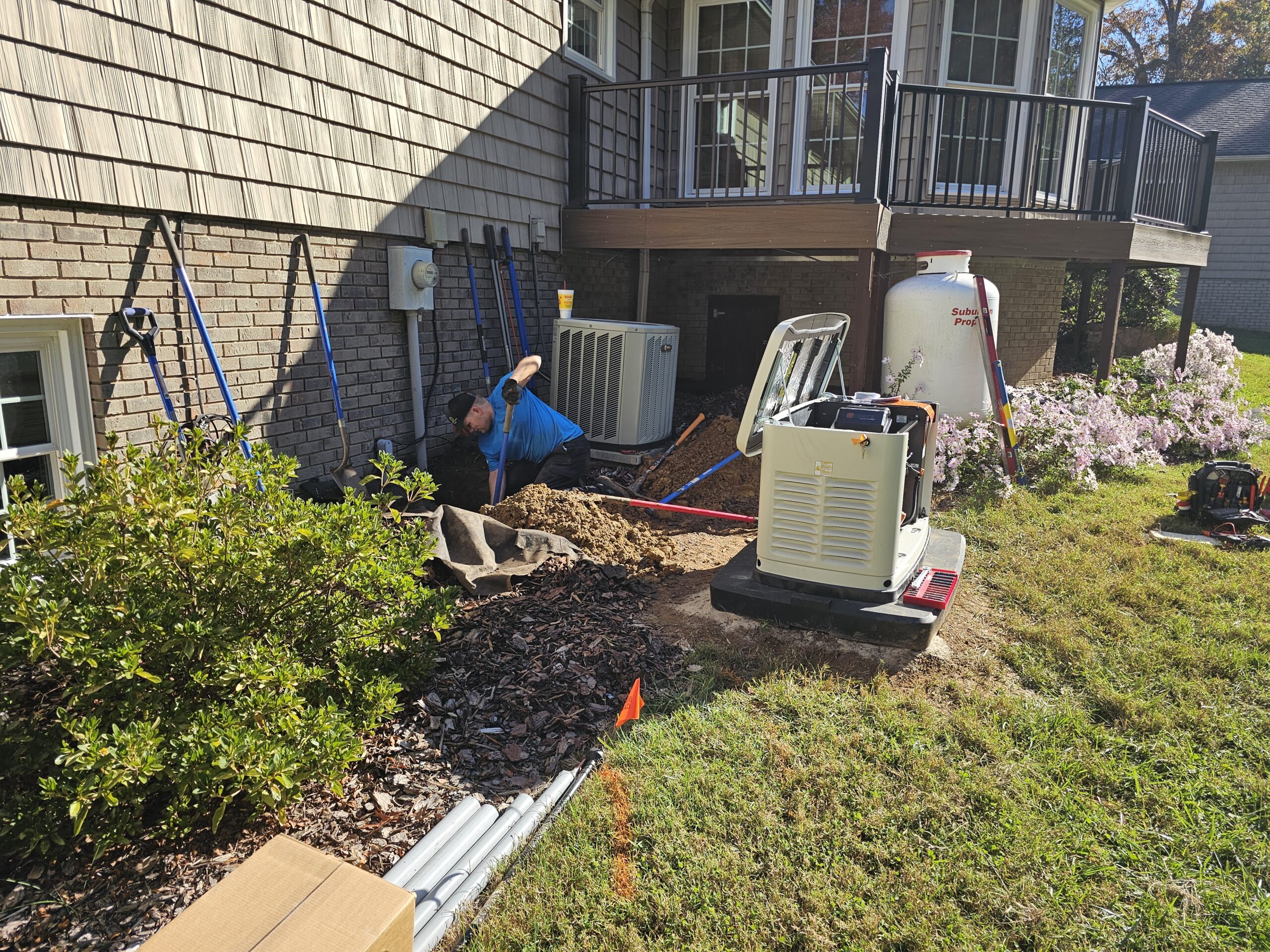A worker uses a shovel to dig a trench beside a house near an HVAC unit; tools and equipment are scattered around the work area.