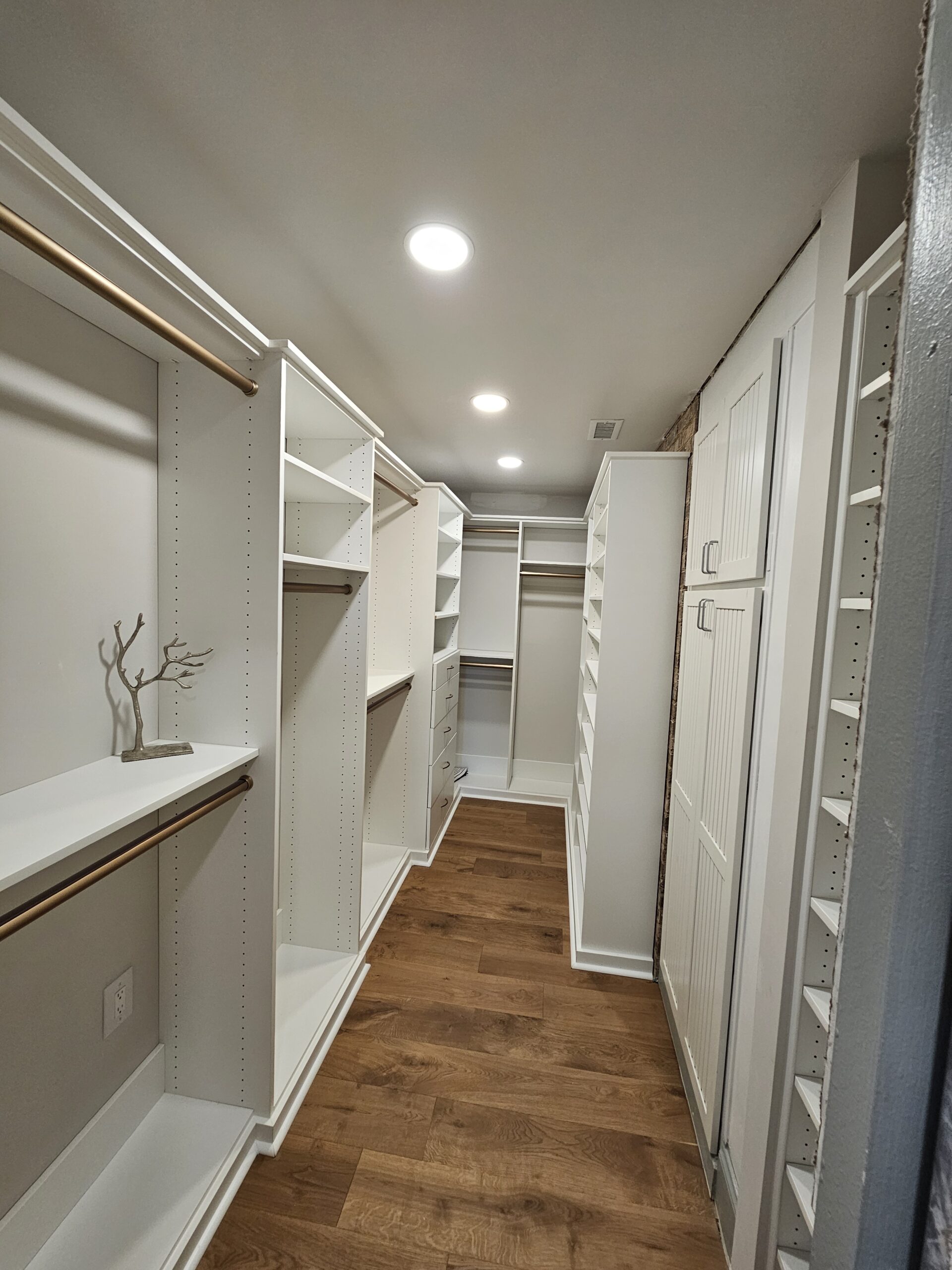 A walk-in closet with white shelves, cabinets, hanging rods, and wooden flooring, illuminated by recessed ceiling lights.