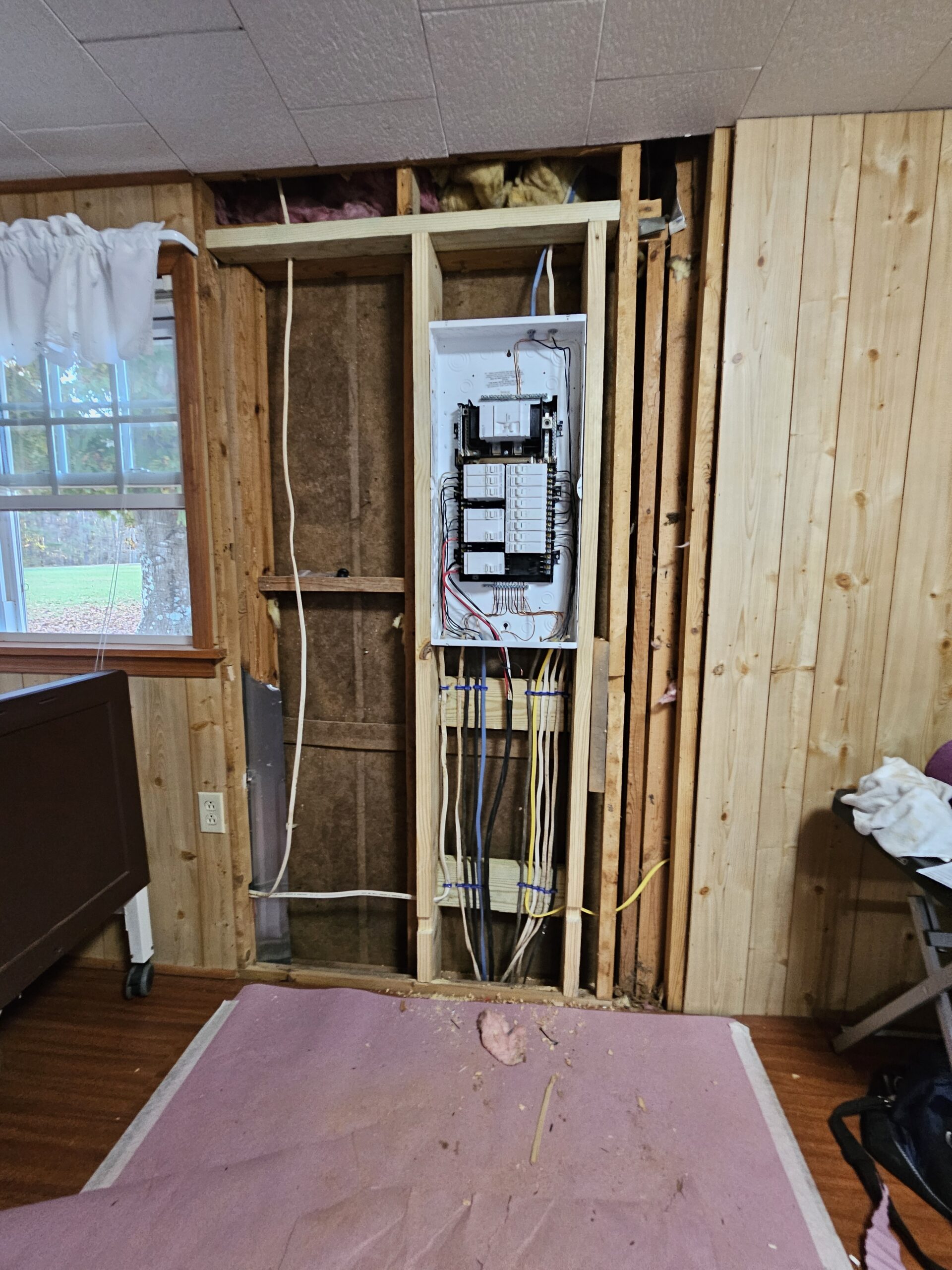 An open wall section reveals a residential electrical panel with exposed wiring in a room with wood paneling and a pink drop cloth on the floor.